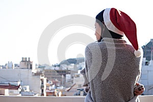 Back view of a young female wearing a santa claus hat and a winter blanket looking at the city