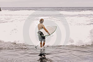 Back view of young fit surfer man with surfboard runs into ocean or sea with big waves for surfing. Concept of extreme