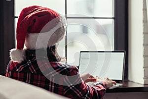 Back view of young freelancer woman in a red santa claus christmas hat sitting near window and working on laptop. Home work