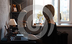 Back view of young girl with blonde braid, working home at graphic tablet on laptop. Looking at window.