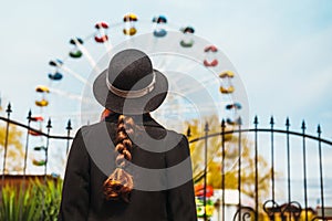 Back view of a young girl in hat standing in front of the ferris wheel at the amusement park