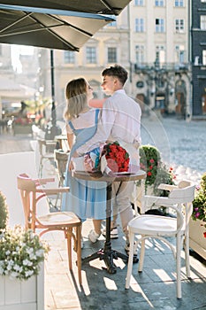 Back view of young happy couple, embracing and looking at each other in light cafe terrace outdoors on a sunny day and