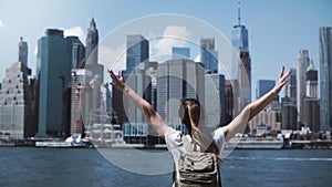 Back view of young happy tourist girl with backpack and arms wide open with joy at famous Manhattan skyline in New York