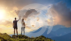 Back view of young hiker couple standing with raised arms holding hands on rocky mountain top enjoying sunset panorama