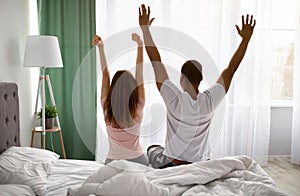 Back view of young interracial couple waking up in morning, stretching on bed at home