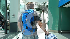 Back of view of young man with backpack pulling suitcase in modern airport terminal. Travelling handsome guy walking