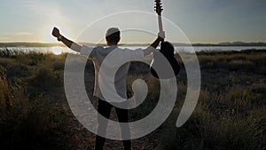 Back view of a young man guitarist raising arms up facing sunset while holding an acoustic guitar - slow-motion