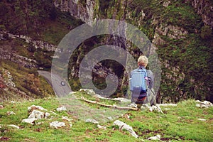 Back view Young man hiker with backpack sitting on mountain`s top with spruce rocks landscape and car road background. Travel