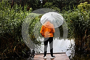 Back view of a young man in an orange raincoat with an umbrella standing on a wooden pier near a lake