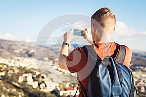 Back view of a young man tourist with backpack taking photo of landscape with smartphone