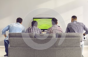 Back view of young men sitting on comfortable sofa and watching football match on TV