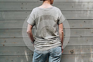 Back view. Young millennial woman dressed in gray t-shirt is stands against gray wood wall.