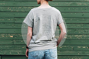 Back view. Young millennial woman dressed in gray t-shirt is stands against green wood wall.