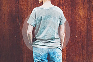 Back view. Young millennial woman dressed in gray t-shirt is stands against wall of rusty metal.