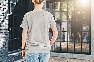 Back view. Young millennial woman dressed in gray t-shirt is stands on city street. Mock up.