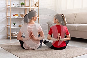 Back view of young mother and her teen daughter doing yoga at home, sitting in lotus pose, making namaste gesture