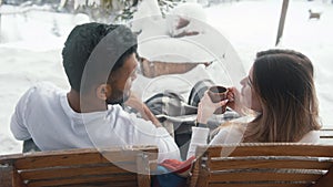 Back view. Young multiracial couple drinking hot chocolate and flirting while sitting on the bench outdoors on