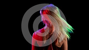 Back view of a young pretty blonde flirtatiously waving her hair. Girl model posing on black studio background with neon