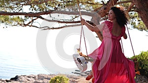 Back View at Young Resting woman in beautiful pink long dress swinging on a swing near the sea