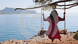 Back View at Young Resting woman in beautiful pink long dress swinging on a swing near the sea