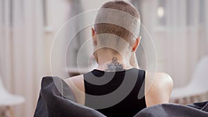 Back view of young slim Caucasian woman having cancer with tattoo on neck sitting indoors thinking. Hairless lady