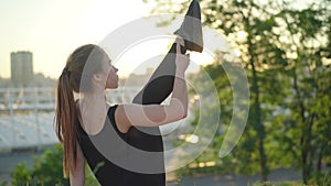 Back view of young sportswoman sitting outdoors at sunset and raising leg. Slim flexible female Caucasian gymnast doing