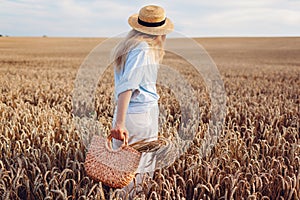 Back view of young stylish woman in hat walking in summer field holding straw handbag with wheat bundle
