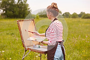 Back view of young woman artist standing in front of sketchbook with brush and palette of colours, paints picture in nature in