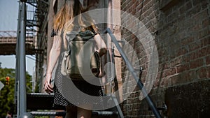 Back view of young woman with backpack and long hair walking up old metal stairs to enter red brick building slow motion