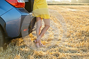 Back view of young woman driver slim legs in short yellow summer dress standing near her car on dry straw field