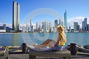 Back view of young woman in hat relaxing sitting on bench enjoy cityscape in Balneario Camboriu, Santa Catarina, Brazil