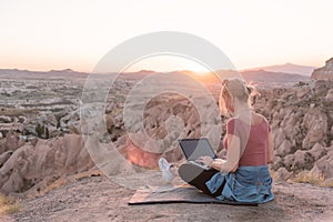 Back view of young woman with laptop sitting on the edge of canyon valley