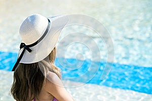 Back view of young woman with long hair wearing straw hat relaxing in summer near swimming pool with blue water on a sunny day