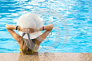 Back view of young woman with long hair wearing yellow straw hat relaxing in warm summer swimming pool with blue water on a sunny