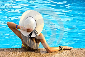 Back view of young woman with long hair wearing yellow straw hat relaxing in warm summer swimming pool with blue water on a sunny