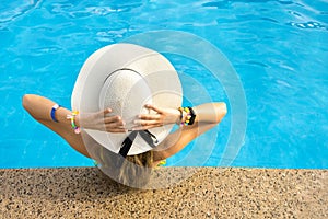 Back view of young woman with long hair wearing yellow straw hat relaxing in warm summer swimming pool with blue water on a sunny