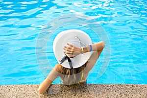 Back view of young woman with long hair wearing yellow straw hat relaxing in warm summer swimming pool with blue water on a sunny