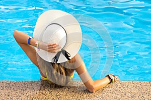 Back view of young woman with long hair wearing yellow straw hat relaxing in warm summer swimming pool with blue water on a sunny