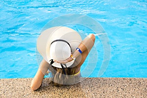 Back view of young woman with long hair wearing yellow straw hat relaxing in warm summer swimming pool with blue water on a sunny