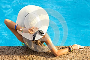 Back view of young woman with long hair wearing yellow straw hat relaxing in warm summer swimming pool with blue water on a sunny