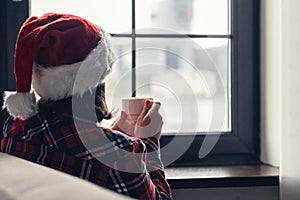 Back view of young woman in a red santa claus christmas hat sitting near window, having breakfast with cup of coffee. Concept.
