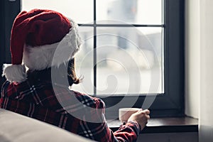 Back view of young woman in a red santa claus christmas hat sitting near window, having breakfast with cup of coffee. Concept.