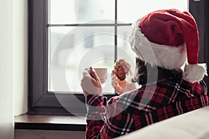 Back view of young woman in a red santa claus christmas hat sitting near window, having breakfast with cup of coffee and croissant