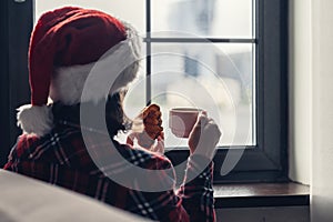 Back view of young woman in a red santa claus christmas hat sitting near window, having breakfast with cup of coffee and croissant