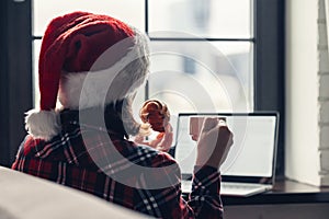 Back view of young woman in a red santa claus christmas hat sitting near window with laptop, having breakfast with cup of coffee