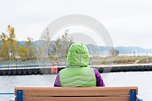 Back view of a young woman sitting alone on the wooden bench in front of the lake. Loneliness concept
