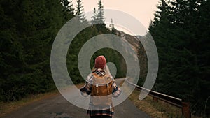 Back view of the young woman traveller walking in middle of empty road in mountains, carrying backpack, enjoying of the