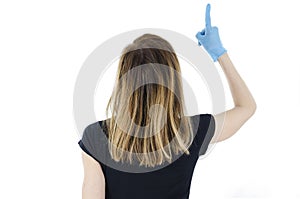 Back view of young woman with white face mask and blue gloves pointing finger up. Rear view of woman pointing at wall.