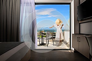 Back view of young woman in white summer dress and straw hat on open balcony terrace of resort hotel room with sea view landscape