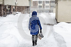 Back of woman in dawn jacket walking through city street during heavy snowfall and blizzard in winter. Bad weather forecast.